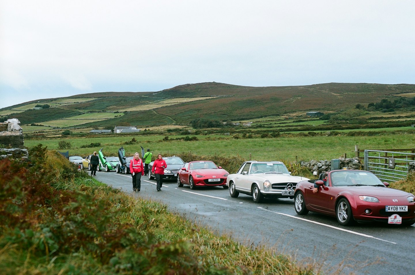 Classic and sports cars on a wet, winding rural road.