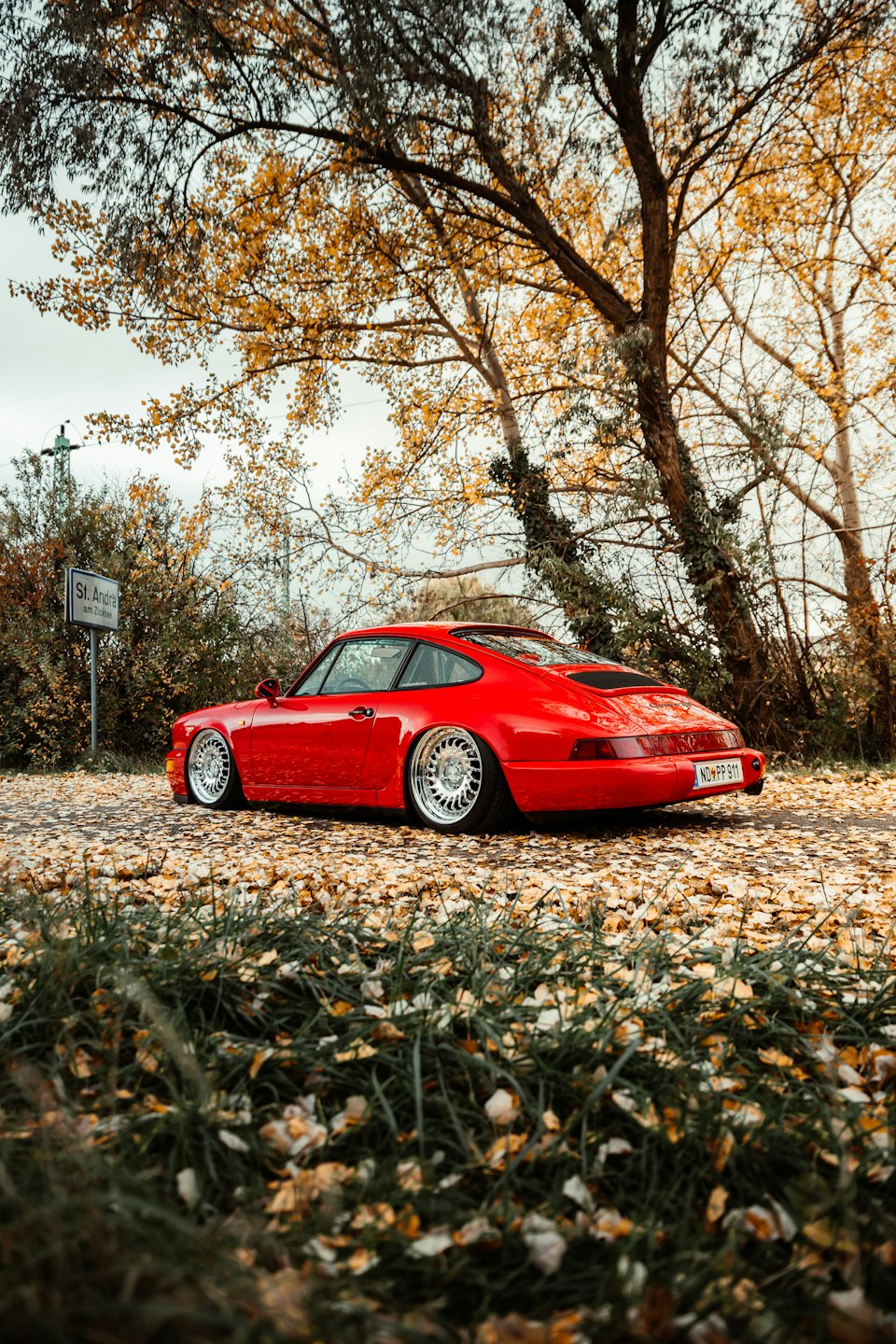 A classic red sports car on a roadside covered in autumn leaves.