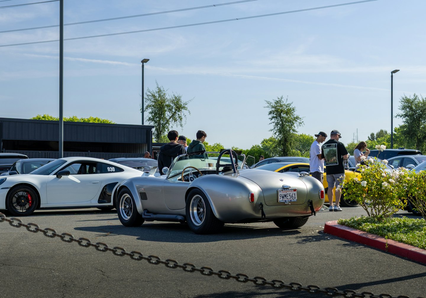 People admire classic and luxury cars in a sunny parking lot.